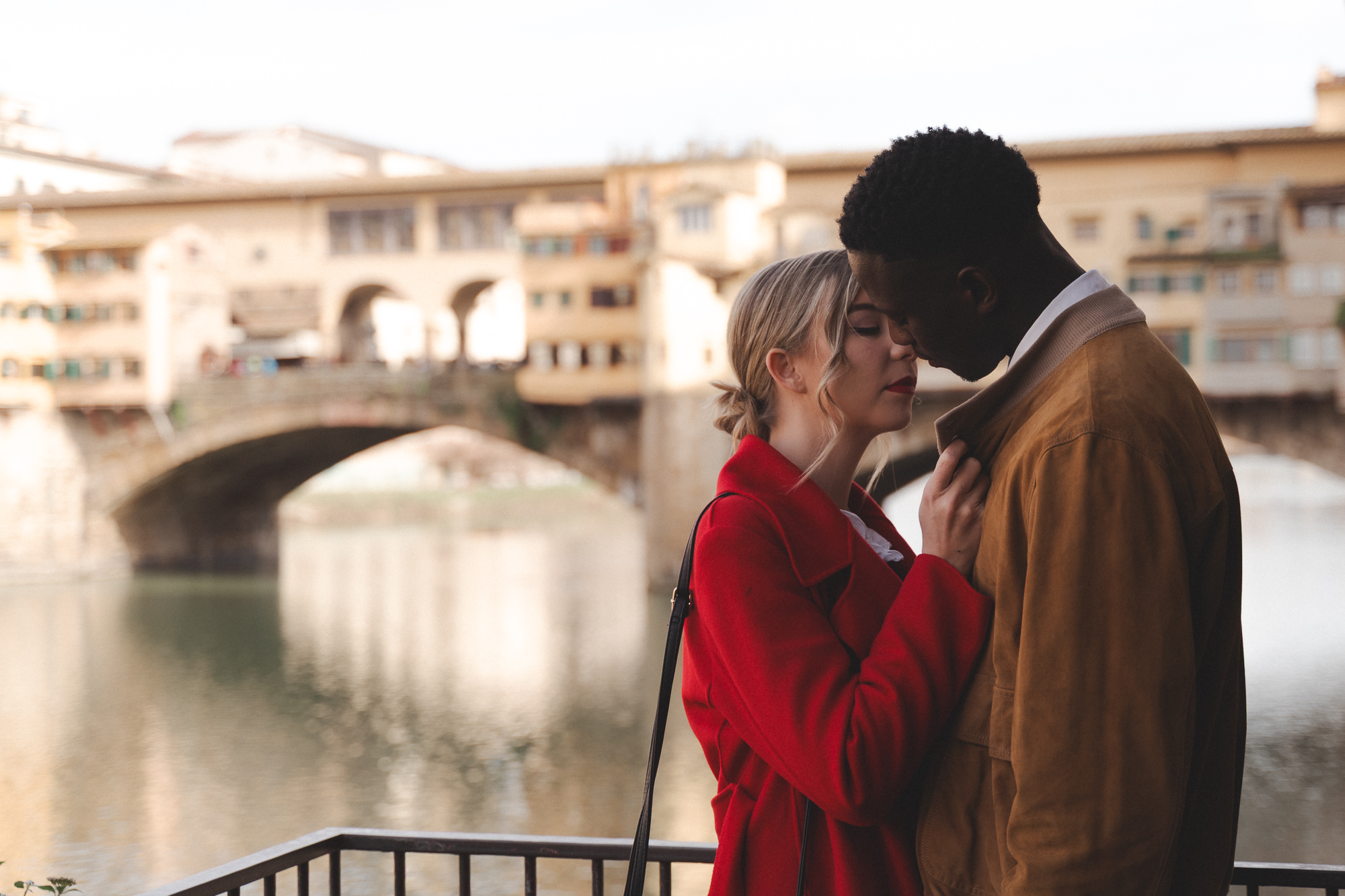 Couple embracing in Florence wearing warm autumn colors like red and tan near the Arno river.