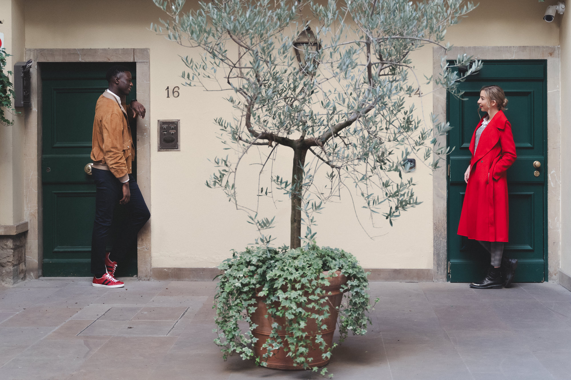 Wide shot of a couple standing at opposite green doors in a courtyard with an olive tree.