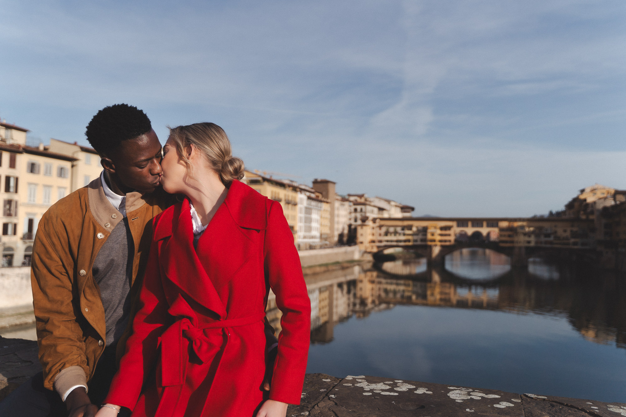Couple kissing on a bridge with the city reflected in the water below.