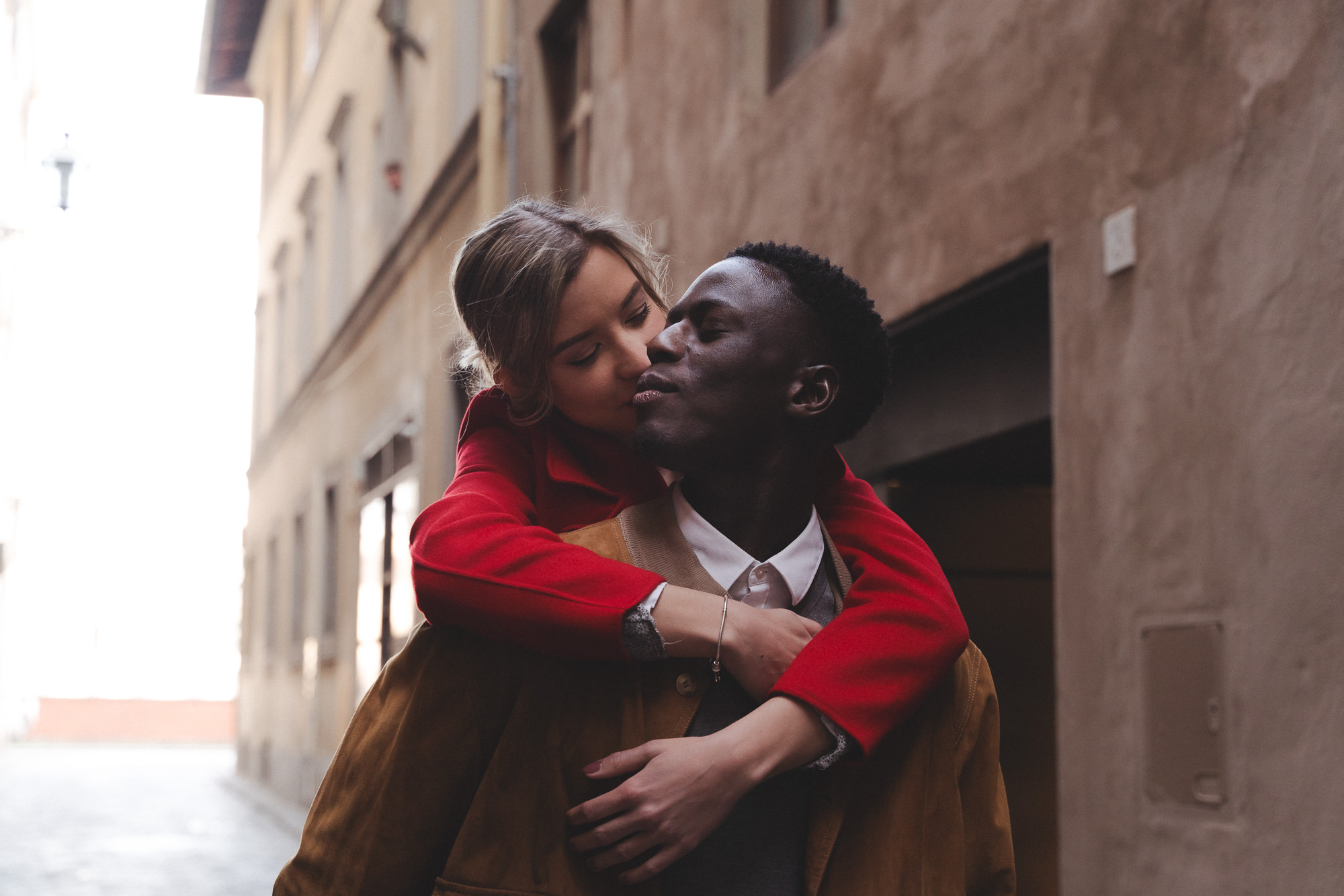 Piggyback ride in a sunlit alley with the couple sharing a kiss.