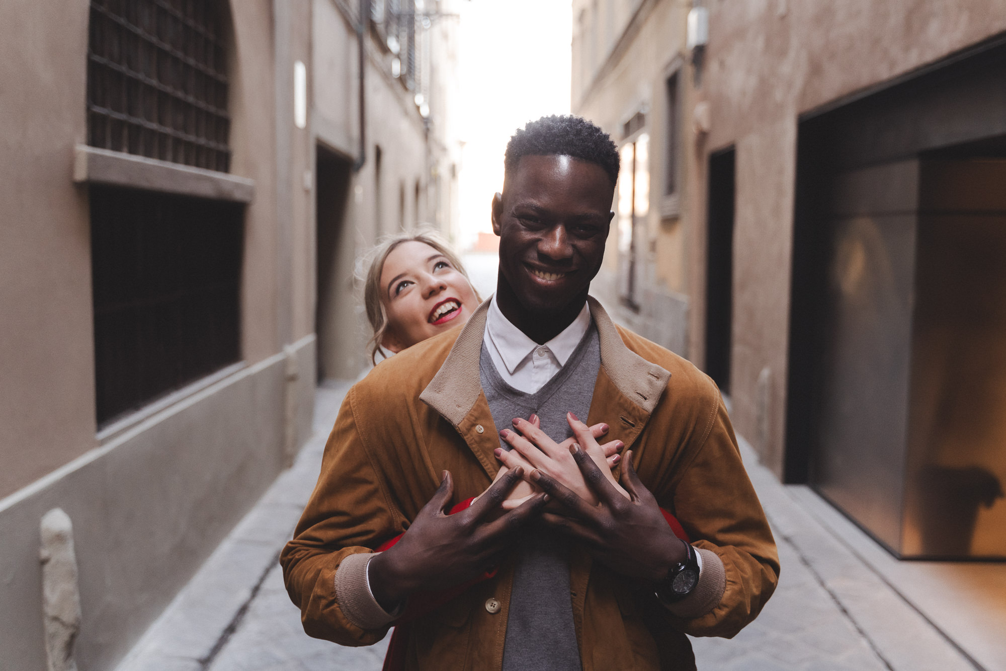Groom hugging bride from behind, both smiling at the camera in an alley