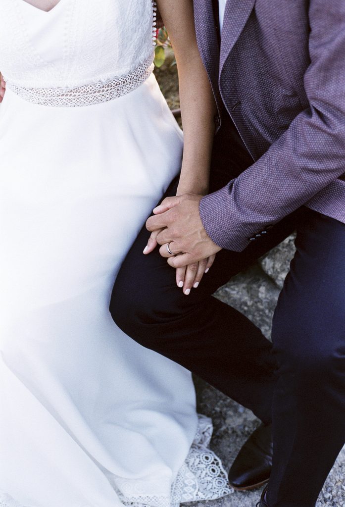 Close-up of a seated couple holding hands, with a white lace wedding dress and a mauve jacket visible.