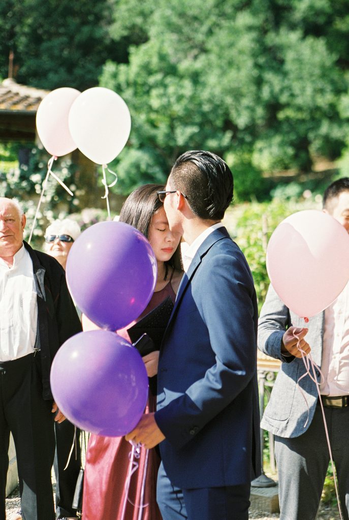 Wedding guest in a blue suit leaning close to a woman among pastel and purple balloons during an outdoor celebration.