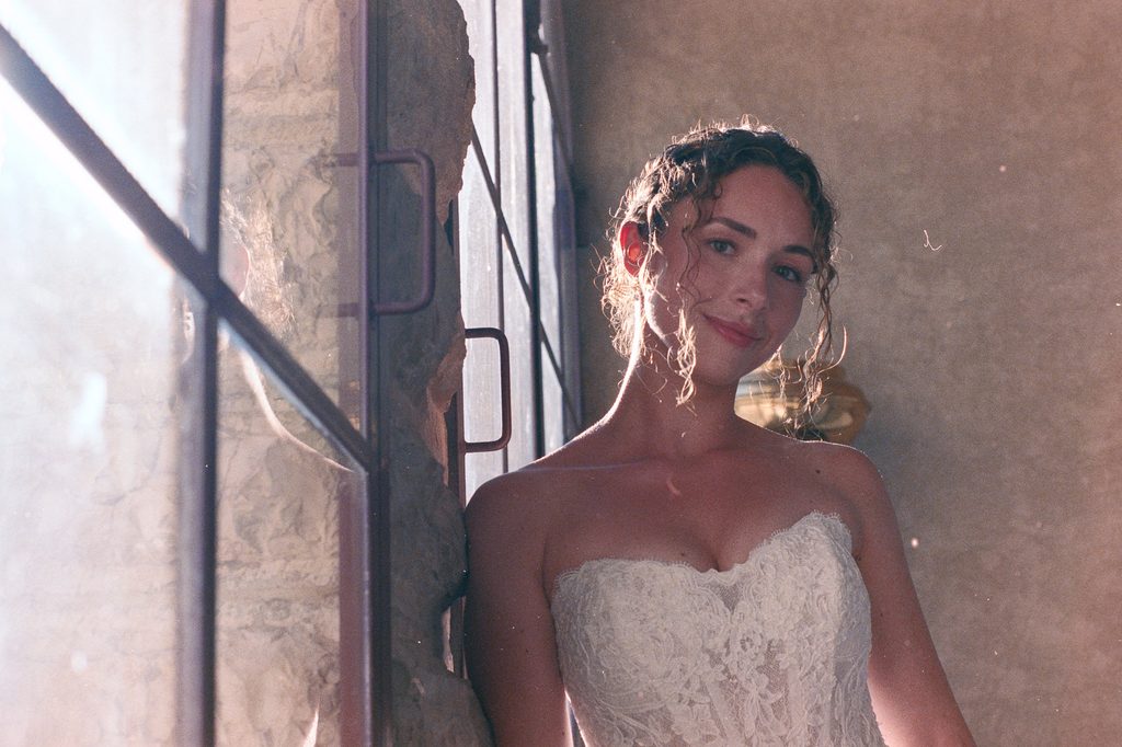 Bride in a lace wedding dress standing beside a window, softly backlit by warm sunlight, with her reflection visible in the glass.